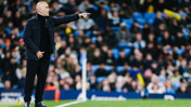 A bald coach in a dark jacket gestures and points from the sideline during a game with a blurred crowd in the background.