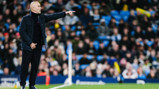 A bald coach in a dark jacket gestures and points from the sideline during a game with a blurred crowd in the background.
