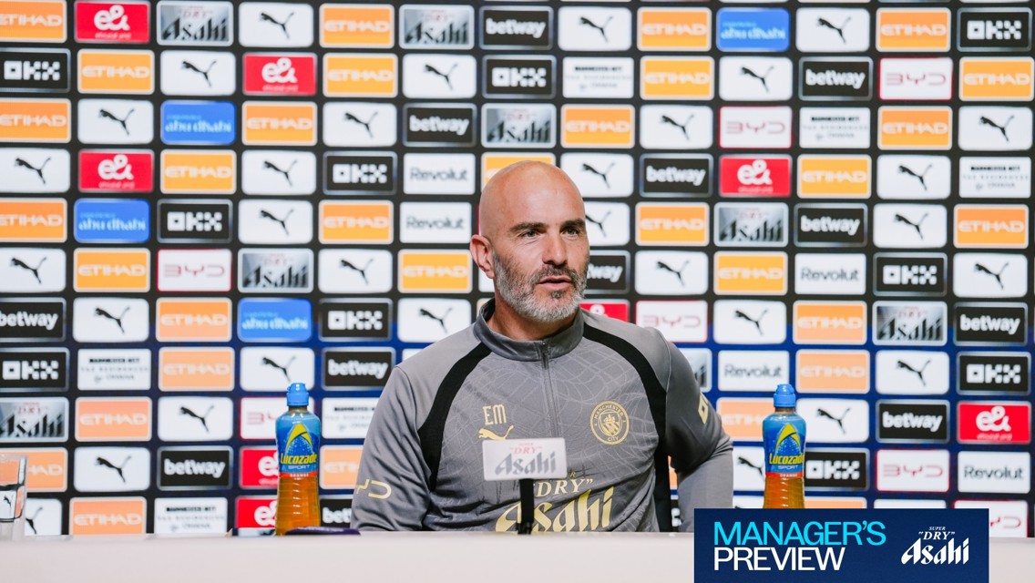 A person in a Manchester City tracksuit sits at a press conference table with branding logos behind them, including Puma and Etihad. There are Lucozade bottles on the table.