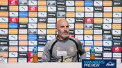 A person in a Manchester City tracksuit sits at a press conference table with branding logos behind them, including Puma and Etihad. There are Lucozade bottles on the table.