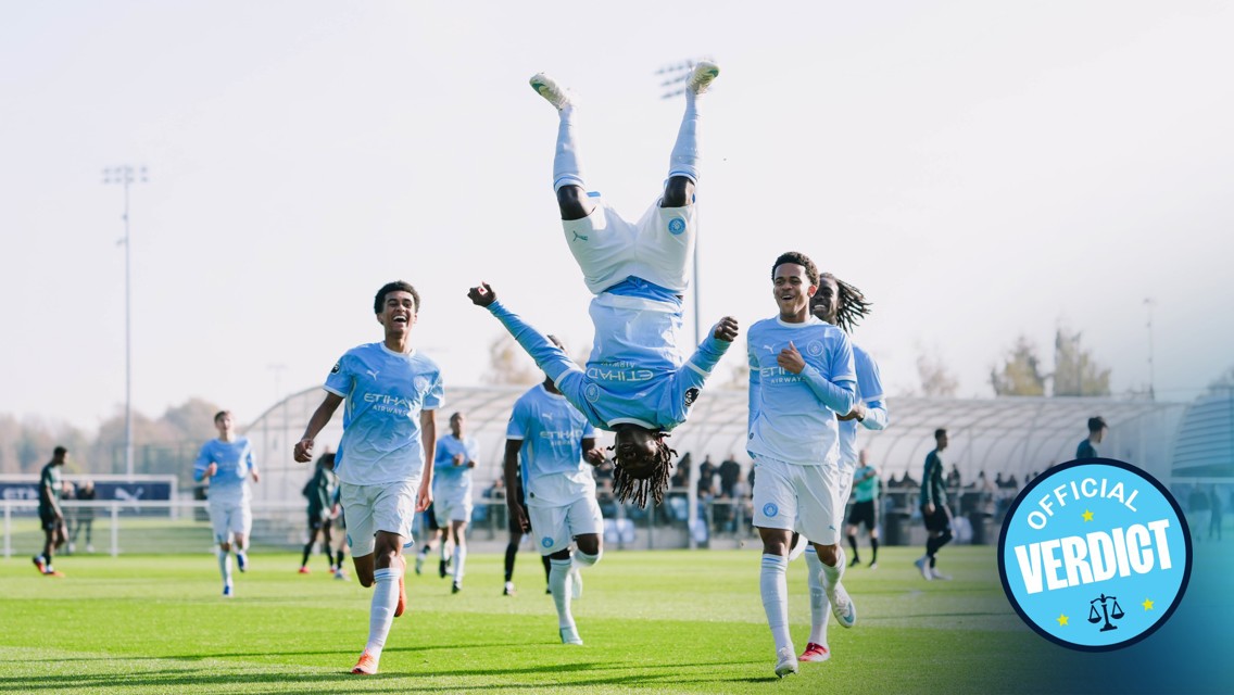Manchester City youth team players celebrate a goal on a soccer field, with one player performing a flip in mid-air. The team wears light blue uniforms, and the setting appears to be a training ground.