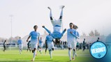 Manchester City youth team players celebrate a goal on a soccer field, with one player performing a flip in mid-air. The team wears light blue uniforms, and the setting appears to be a training ground.