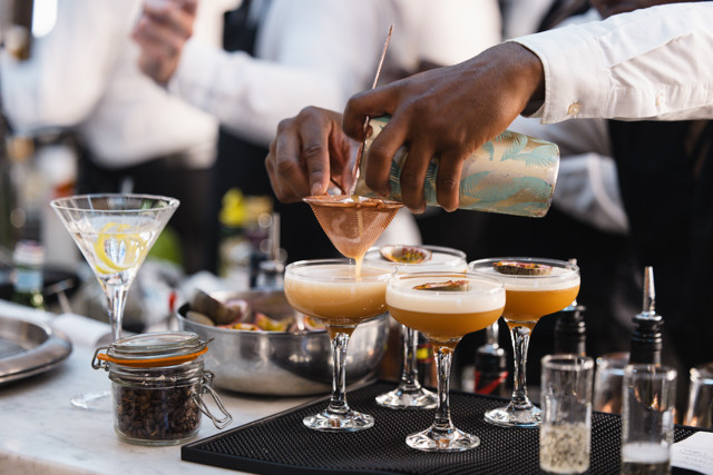 A bartender is seen pouring drinks through a strainer into coupe glasses at a bar, with a cocktail shaker and martini glass visible in the background.
