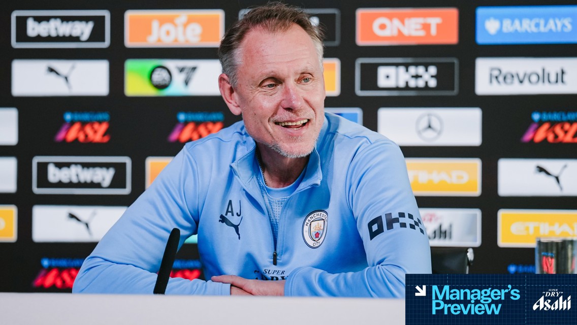 A person with initials AJ, wearing a Manchester City tracksuit, is speaking at a press conference. Sponsor logos are displayed in the background.