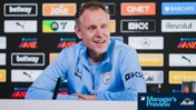 A person with initials AJ, wearing a Manchester City tracksuit, is speaking at a press conference. Sponsor logos are displayed in the background.