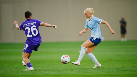 Two female soccer players are competing for control of the ball during a match. One player is wearing a purple jersey with the number 26, and the other wears a light blue kit.