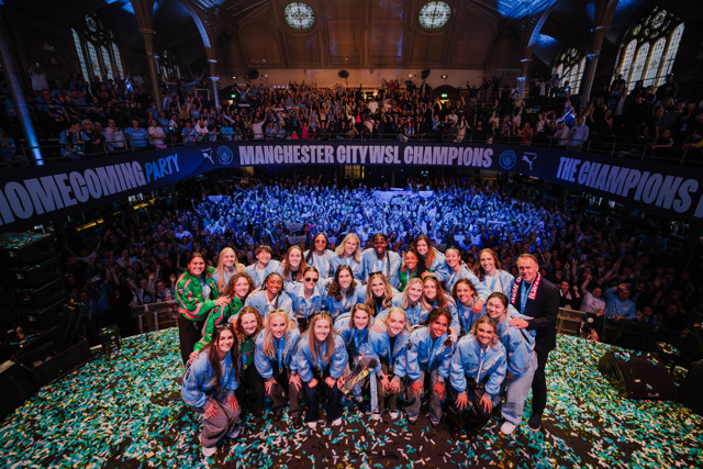 Manchester City celebrates WSL championship with team on stage, confetti, and large crowd in venue adorned with banners.