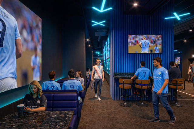Fans watching a football match in a Manchester City-themed lounge with screens showing players wearing City jerseys.