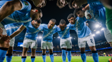 Manchester City players in a huddle on the field at night with stadium lights illuminating the scene.