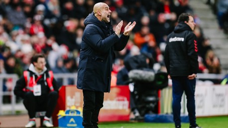 A football coach wearing a dark coat claps during a match. The venue is crowded with spectators in the background and team staff visible on the sideline.