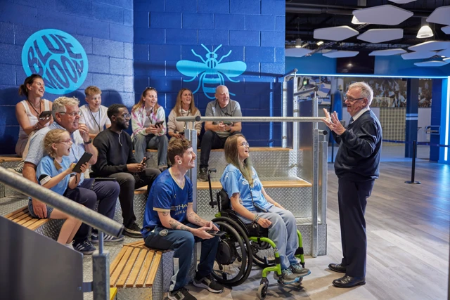 A group of people, including a person in a wheelchair, listening attentively to a tour guide inside the Manchester City stadium. The background features a blue bee logo and 'Blue Moon' sign on the wall.