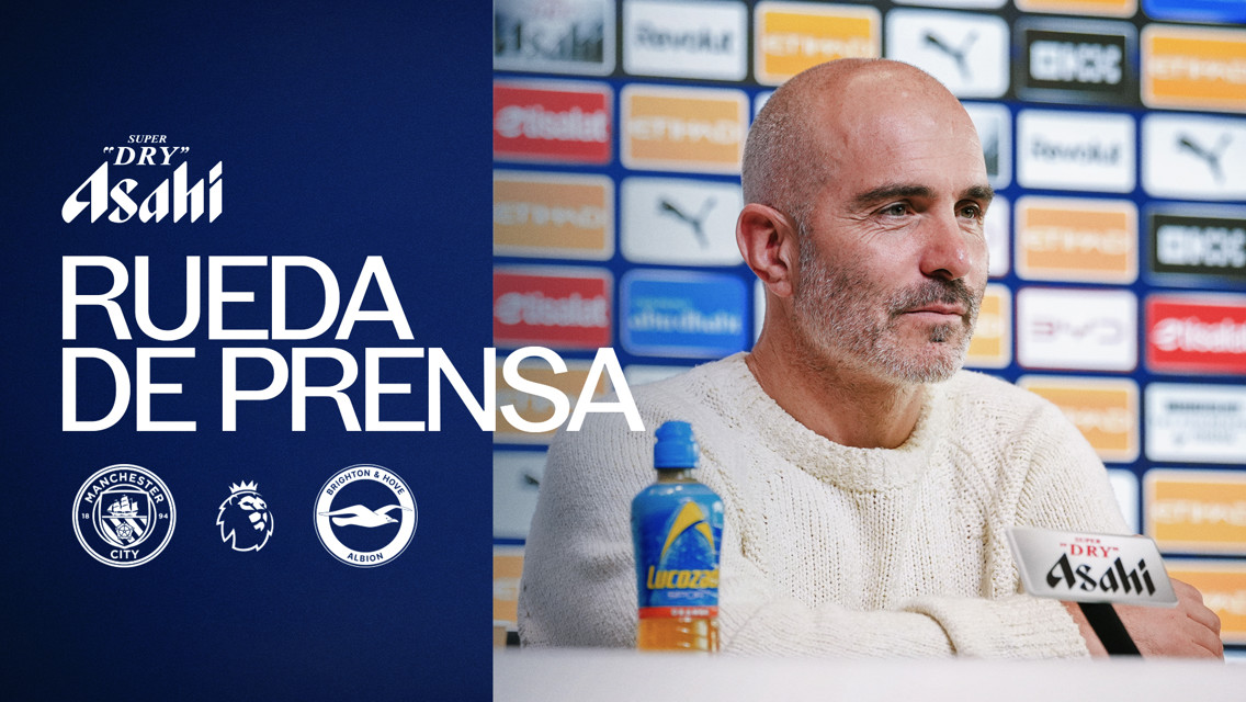 A man in a gray sweater speaks at a press conference with branding from Manchester City and Brighton & Hove Albion, sponsored by Asahi. Puma and other logos are visible in the background.