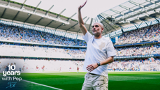 A man stands on a football pitch at Etihad Stadium celebrating 10 years with Pep. The stadium is filled with spectators.