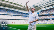 A man stands on a football pitch at Etihad Stadium celebrating 10 years with Pep. The stadium is filled with spectators.
