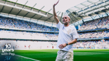 A man stands on a football pitch at Etihad Stadium celebrating 10 years with Pep. The stadium is filled with spectators.
