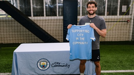 A person holds a Manchester City jersey with 'Supporting City in the Community' printed on it, standing indoors on a turf field next to a table with the Manchester City logo.