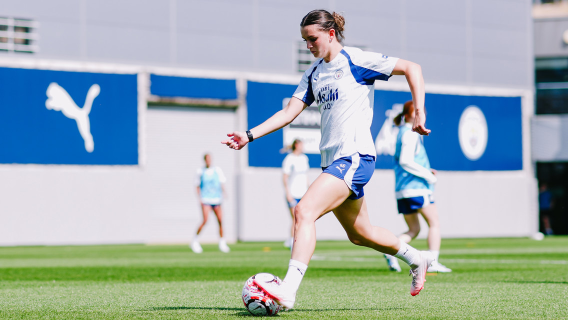 A woman in a Man City training kit is playing football on a field, with other players visible in the background.