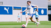 A woman in a Man City training kit is playing football on a field, with other players visible in the background.