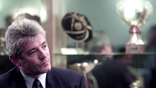 A man in a suit with a serious expression is looking to the side. Behind him, there's a showcase with awards and a cap.