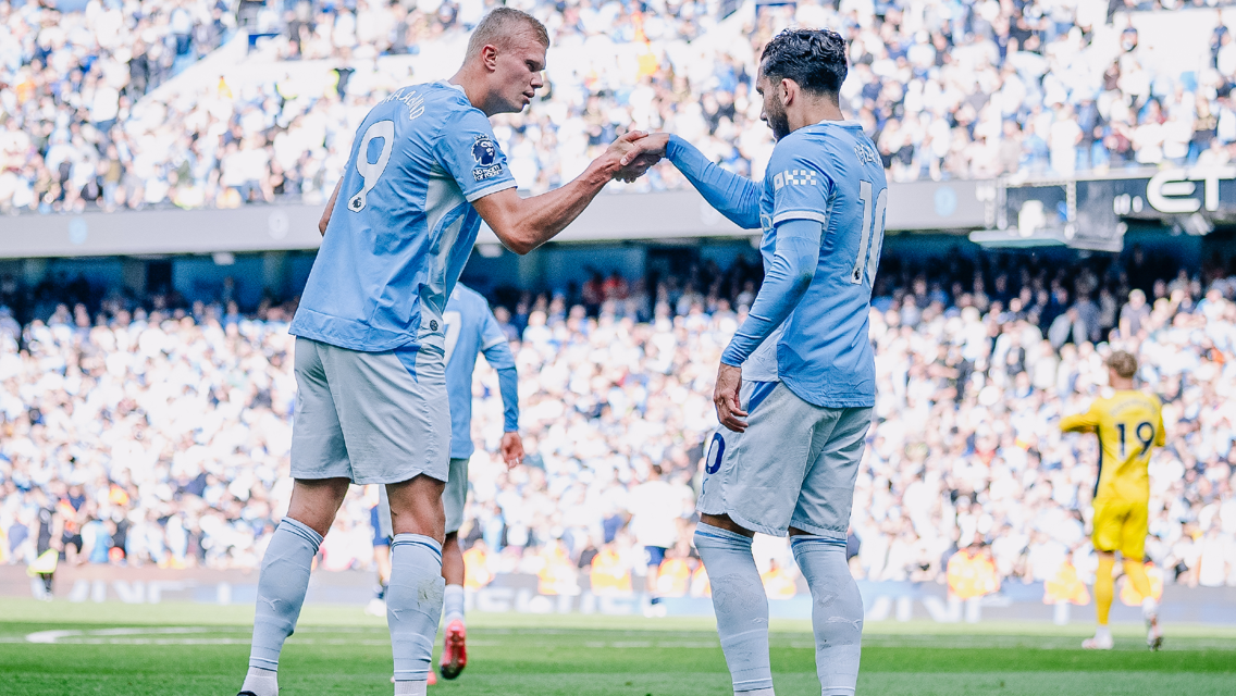 Two Manchester City players in light blue jerseys fist bump on the field. One player wears number 9 and the other number 20. The crowd is visible in the background.
