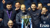 Group of individuals celebrating a trophy win. The trophy has 'Premier League' ribbons. Text mentioning 'Welcome back Enzo' and 'Manchester City'.