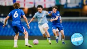 A soccer match between Manchester City and Chelsea players, with one player in Manchester City kit and two in Chelsea kit, engaging in a goal attempt.