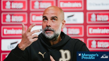 A person with a blurred face at a press conference with a red Emirates FA Cup background and a Manager's Preview overlay.