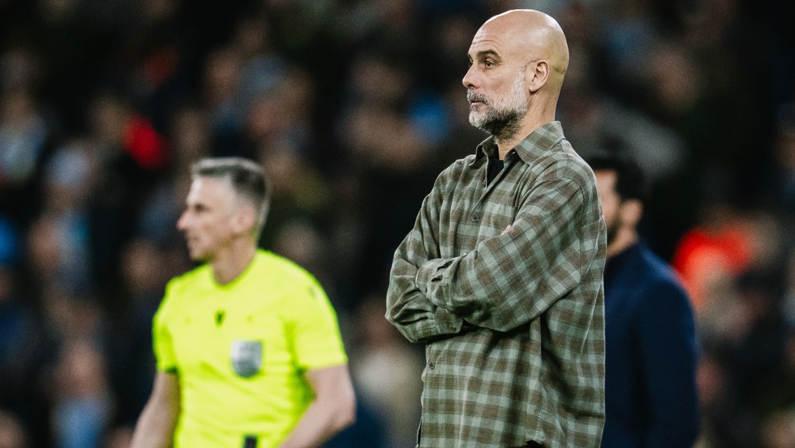 A bald man wearing a plaid shirt stands with arms crossed on the sideline of a sports field. A person in a bright yellow outfit is in the background.