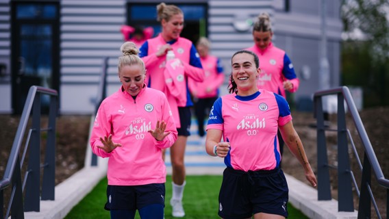 Group of people wearing pink Manchester City training tops walk down stairs outside a building.