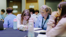 A group of students sitting at tables in a classroom, engaged in writing and discussion, with notebooks and water bottles on the tables.