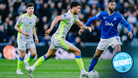 Football players from Manchester City and Everton competing for the ball during a match. Manchester City player in grey and fluorescent green kit, Everton player in blue kit.