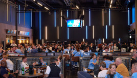 A bustling bar filled with football fans wearing team jerseys, watching a game on a screen, and socializing. The atmosphere is lively with blue lights illuminating the interior.