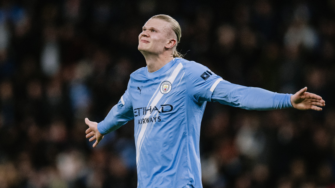 A Manchester City player in a blue kit, with outstretched arms celebrating a goal. The player's face is blurred. The Etihad Airways logo is visible on the jersey.