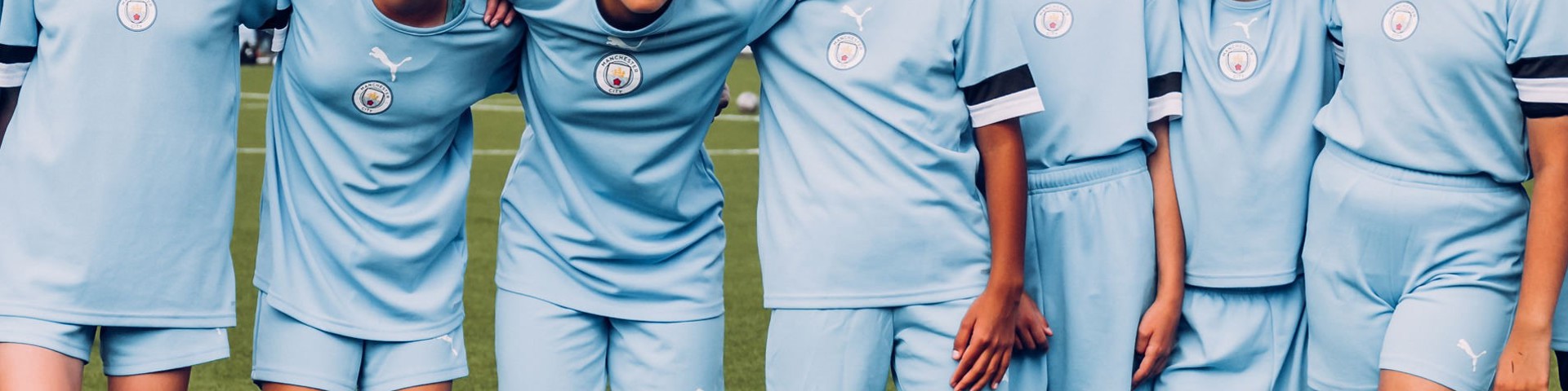 A group of seven children in matching light blue football kits with the Manchester City logo pose for a team photo on a field.
