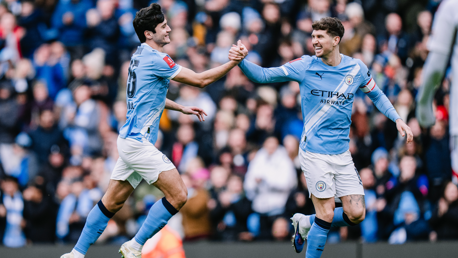 Two Manchester City players wearing light blue jerseys celebrate during a football match with a handshake.