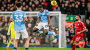 Players from Manchester City and Liverpool compete for the ball during a soccer match. A Manchester City player is jumping to head the ball.