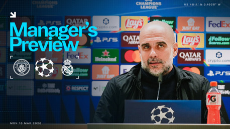 An image showing a blurred manager at a press conference for the UEFA Champions League match between Manchester City and Real Madrid, with logos and sponsor banners in the background.