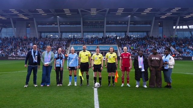 Soccer players, referees, and fans pose on a green field with a stadium in the background.