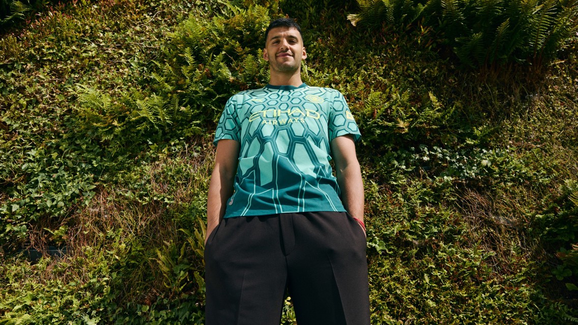 Person wearing a Manchester City shirt with a hexagonal pattern, standing against a wall of greenery.