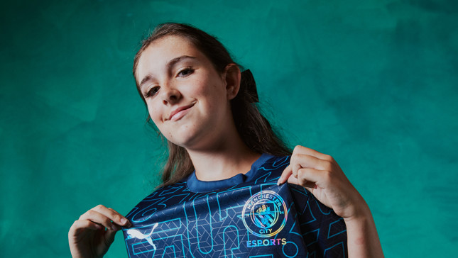 A female with a blurred face is holding a Manchester City Esports jersey in front of a green background.