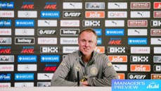 A man in a grey jacket sits at a press conference table with microphones, in front of a backdrop of sponsor logos, including Manchester City Logo and Barclays WSL.