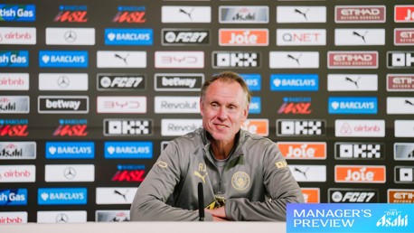A man in a grey jacket sits at a press conference table with microphones, in front of a backdrop of sponsor logos, including Manchester City Logo and Barclays WSL.