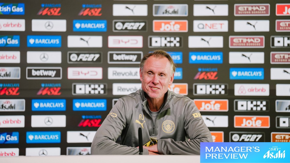 A man in a grey jacket sits at a press conference table with microphones, in front of a backdrop of sponsor logos, including Manchester City Logo and Barclays WSL.