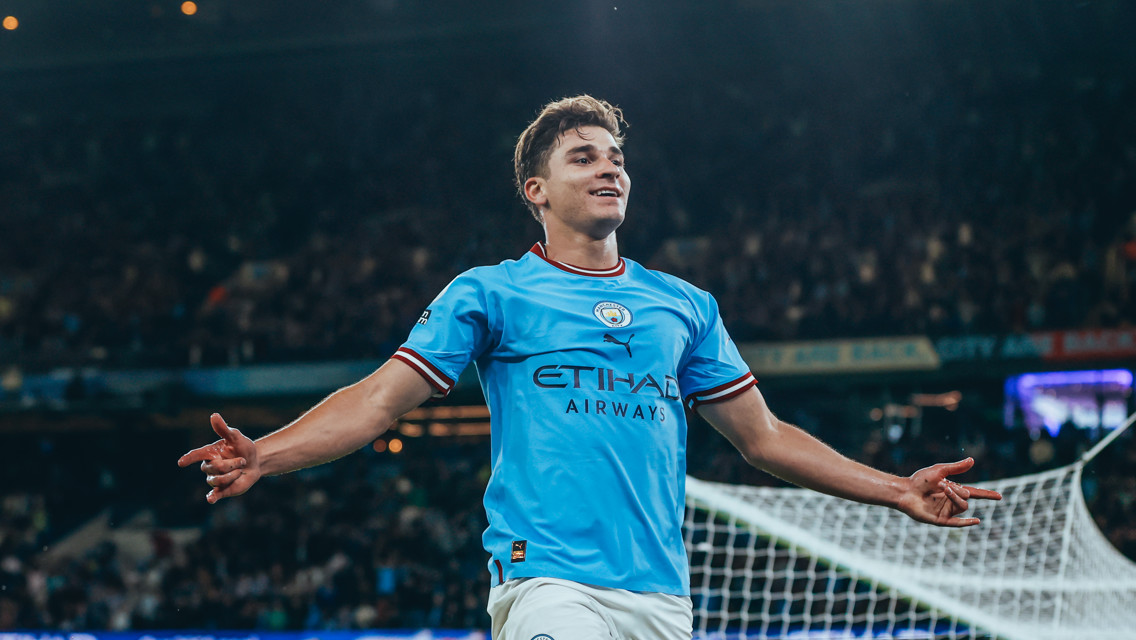 A football player wearing a Manchester City kit celebrates in front of a goal net during a night match.
