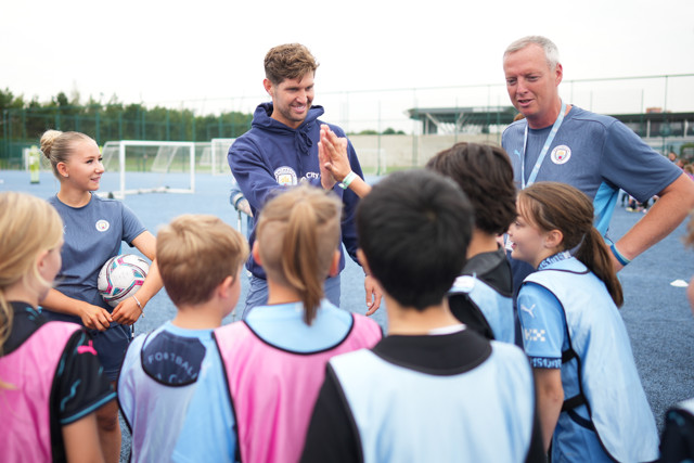 A group of children in light blue vests along with coaches, one holding a soccer ball, at a Manchester City youth training session on a sports field.