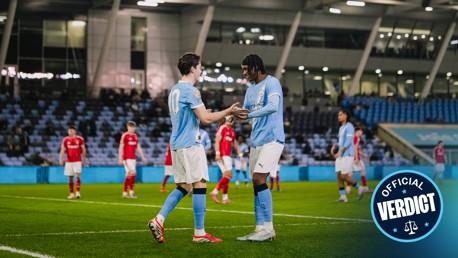 Players wearing Manchester City jerseys embrace on the football field after scoring a goal.