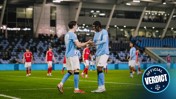 Players wearing Manchester City jerseys embrace on the football field after scoring a goal.