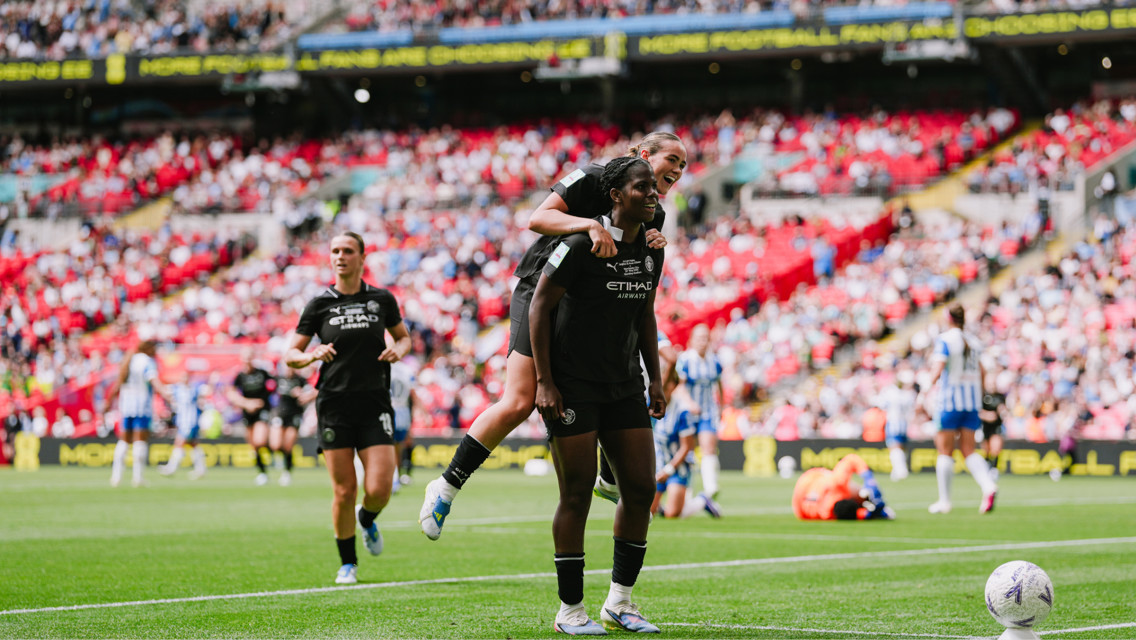A football match scene with players celebrating on the field. One player is carrying another on their back. The team is wearing black kits, branded with Etihad Airways, and playing in a stadium with visible crowd.