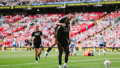A football match scene with players celebrating on the field. One player is carrying another on their back. The team is wearing black kits, branded with Etihad Airways, and playing in a stadium with visible crowd.