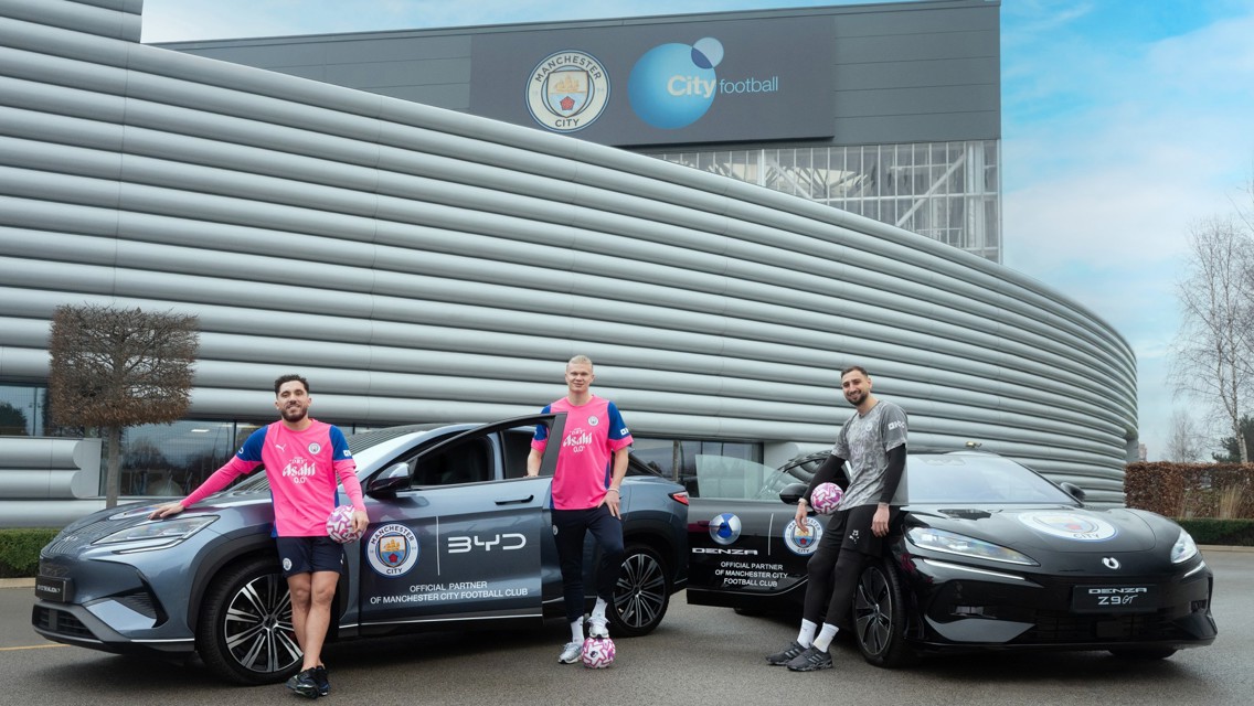 Manchester City players posed in front of electric cars bearing the club's logo, next to a building featuring the Manchester City and City Football Group logos.